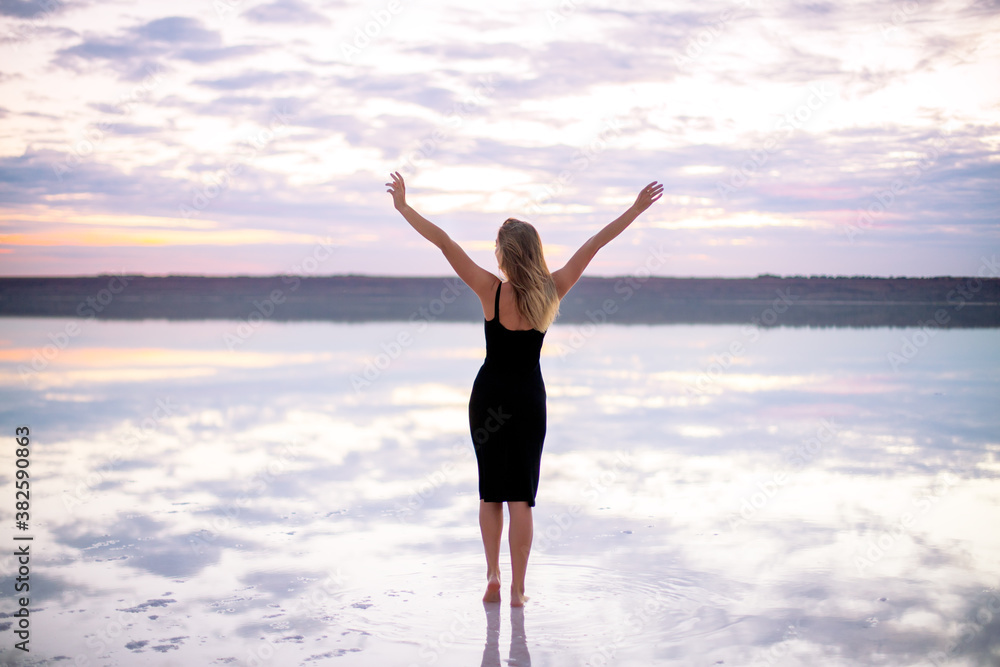 beautiful girl in a black dress on a salty pink lake at a vanilla sunset raised her hands
