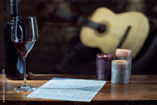 Music notes, wine, candle on wooden table, with a guitar in the background.