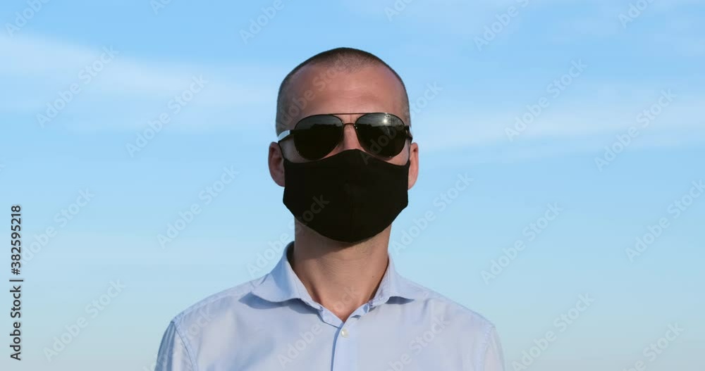 A man with short hair in a black face mask and sunglasses looks into a video camera and holds a smartphone to his ear against a blue sky with clouds. A man with short hair in a black face mask and sunglasses looks into a video camera and holds a smartphone to his ear against a blue sky with clouds.