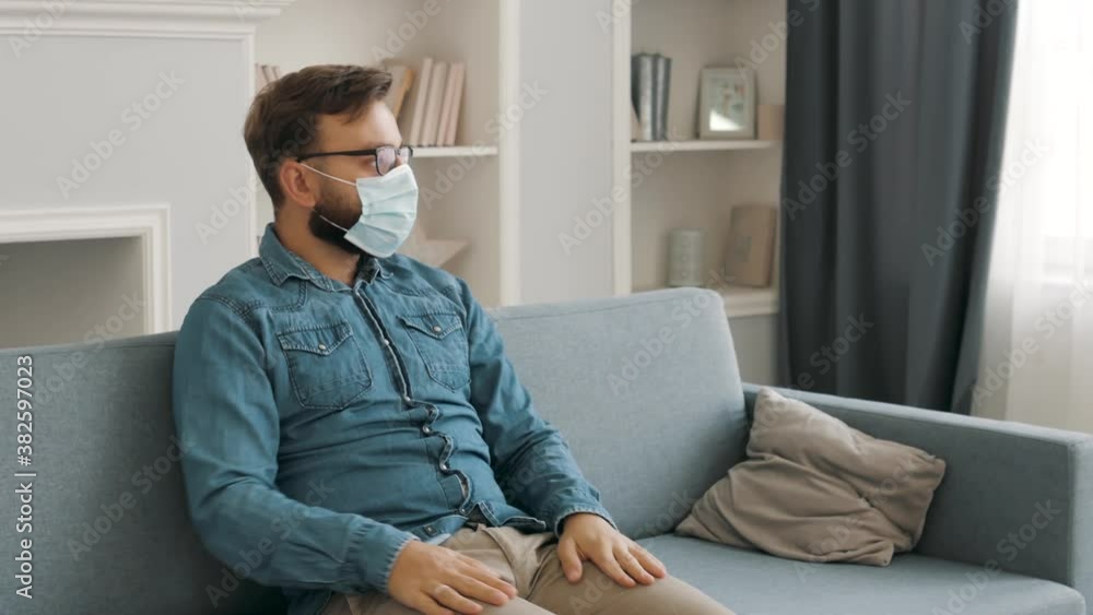 Isolated Sick young caucasian Young man in medical protective mask sitting on a sofa, coughing, sneezing at home. coronavirus, quarantine, pandemic time Isolated Sick young caucasian Young man in medical protective mask sitting on a sofa, coughing, sneezing at home. coronavirus, quarantine, pandemic time