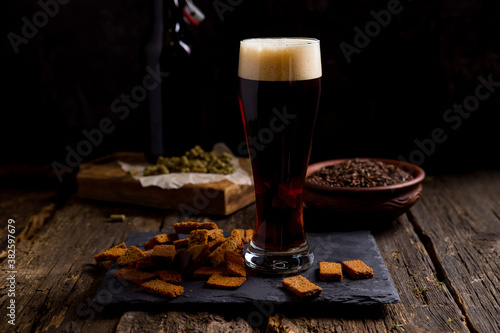 dark beer with foam in a glass. On a dark background bottle and ingredients for beer, malt, hops. Home brewery. Background image