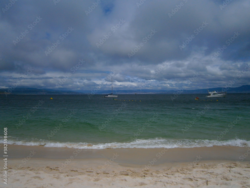 Waves against the sand under a cloudy sky with boats on the ocean