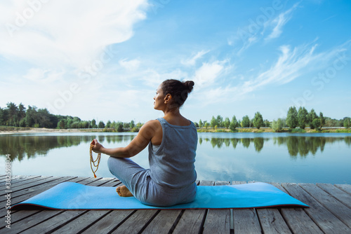 A young woman sits on a mat on a wooden pier in a lotus position with a rosary in her hands. Back view. Meditation, yoga in nature.