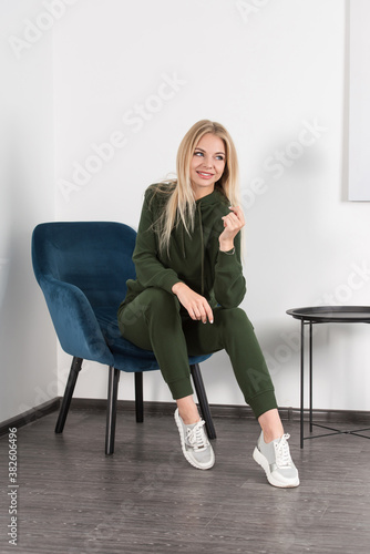 Stylish beautiful young blond woman in a green tracksuit poses near a white wall in the room. Attractive girl model posing in a blue chair.