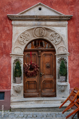 Brown old elegant front wooden carved door with a decorative flower wreath, green plants in a pot, red wall. Cesky Krumlov, South Bohemia, Czech Republic
