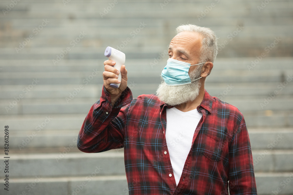 Bearded senior man in face mask holding infrared thermometer a healthy senior man's normal body temperature for coronavirus. Covid-19. Bearded senior man in face mask holding infrared thermometer a healthy senior man's normal body temperature for coronavirus. Covid-19.