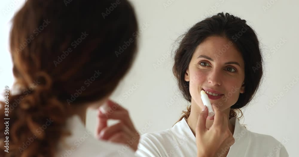 Attractive 30s woman in bathrobe looking in mirror holding lipstick tube apply lips balm. Beauty and make up, decorative cosmetics product advertisement, morning routine self-grooming lip care concept Attractive 30s woman in bathrobe looking in mirror holding lipstick tube apply lips balm. Beauty and make up, decorative cosmetics product advertisement, morning routine self-grooming lip care concept