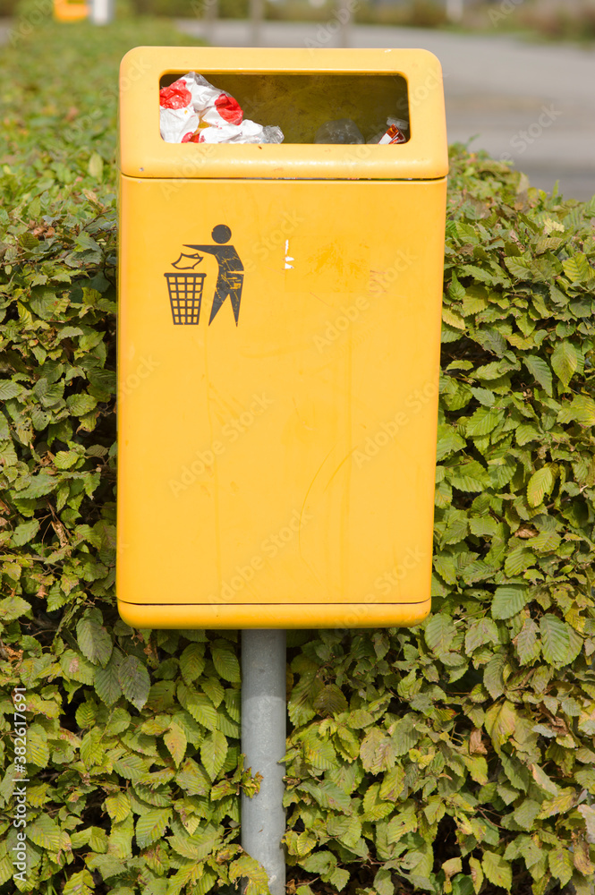 Public yellow waste bin with garbage on the street with green shrubs in the background Public yellow waste bin with garbage on the street with green shrubs in the background