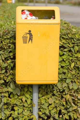 Картината върху платно Public yellow waste bin with garbage on the street with green shrubs in the back