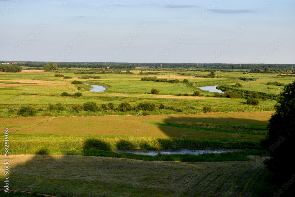View from the top of a tall hill showing a number of vast fields, meadows, and pasturelands, together with a small lake meandering through the area seen on a sunny summer day on a Polish countryside 
