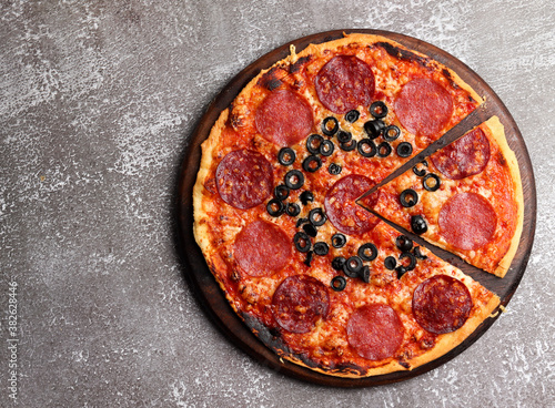 Pizza with pepperoni and olives on a round wooden cutting board on a dark background. Top view, flat lay