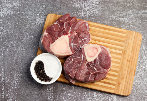 Raw beef shank with salt and pepper on a rectangular cutting wooden board on a dark background. Top view, flat lay