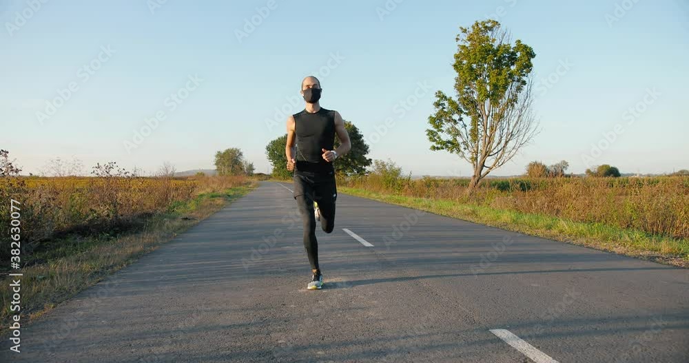 Caucasian handsome strong male jogger running on road in countryside on summer day. Attractive sporty man in mask jogging outside. Pandemic coronavirus concept. Sportsman runner. Caucasian handsome strong male jogger running on road in countryside on summer day. Attractive sporty man in mask jogging outside. Pandemic coronavirus concept. Sportsman runner.