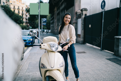 Funny female tourist dressed in casual clothing rejoicing at urban street ready for driving getaway adventure on retro moped bike, toothy Asian hipster girl laughing near vintage rented motorcycle photo