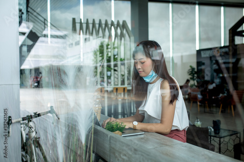 Fototapeta Naklejka Na Ścianę i Meble -  Asian Business Woman Wearing protective Mask working online on laptop at coffee shop in quarantine due to coronavirus.