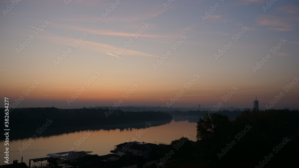 Sunrise Time Lapse on Danube River with Belgrade Serbia Buildings in Background. Blue and Orange Sky with Nature in the Landscape and Trees Reflecting on the Calm Waters of the Danube River.