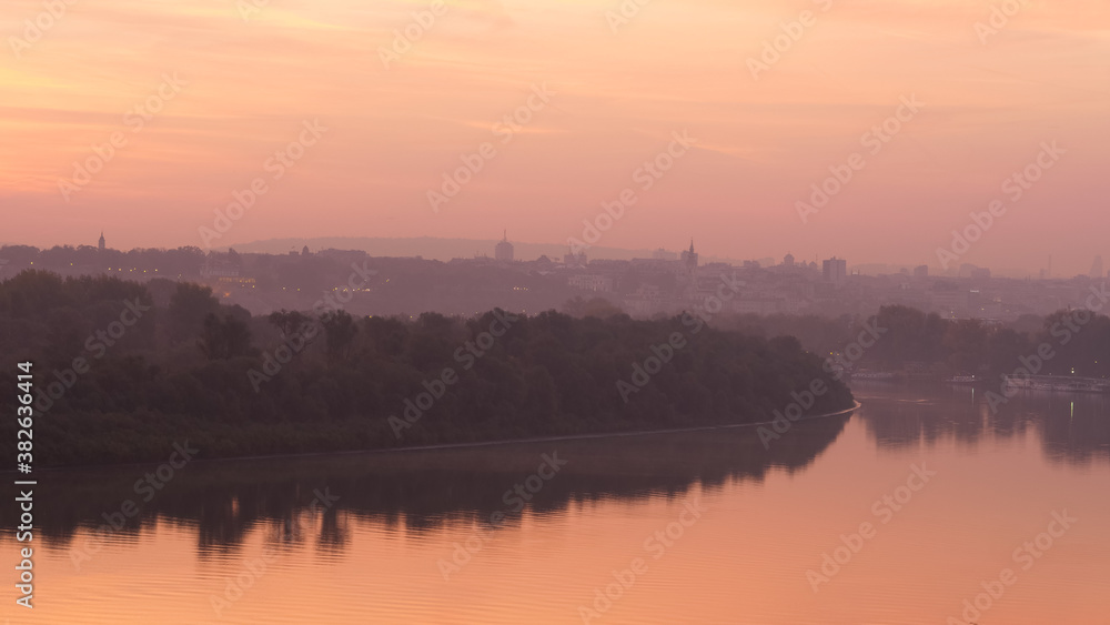 Danube River Bank Landscape with Trees Reflecting on Water at Sunrise in Timelapse with Belgrade Serbia City Downtown with Modern and Historical Buildings of Belgrade City Downtown in the Background. 