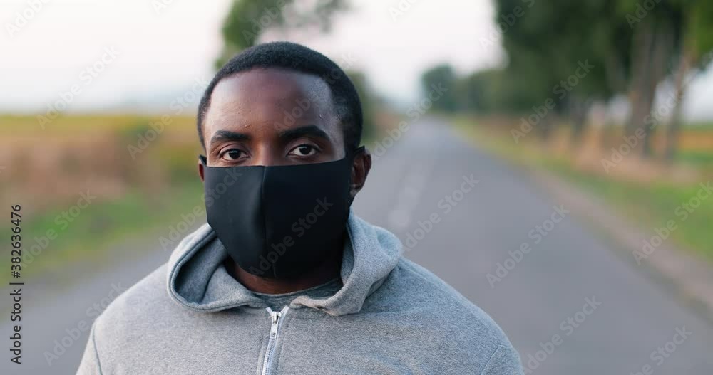 Close up of handsome young African American sportsman in mask in countryside turning face to camera. Portrait of sporty guy jogger standing on road on summer day.