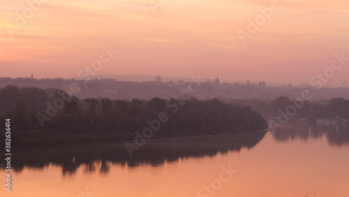 Danube River Bank Landscape with Trees Reflecting on Water at Sunrise in Timelapse with Belgrade Serbia City Downtown with Modern and Historical Buildings of Belgrade City Downtown in the Background. 