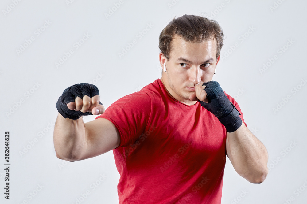 Young man boxes, making hook with right hand. Black bandages, red T-shirt, determined face expression, strong emotions. Concept of blow to kill or hit, copy space on light background. Young man boxes, making hook with right hand. Black bandages, red T-shirt, determined face expression, strong emotions. Concept of blow to kill or hit, copy space on light background.