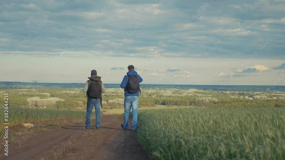 teamwork travel. two man hikers with backpacks walk along a trail next lifestyle to a field with green grass in nature. concept adventure travel hiking healthy. two tourists walk walking talking teamwork travel. two man hikers with backpacks walk along a trail next lifestyle to a field with green grass in nature. concept adventure travel hiking healthy. two tourists walk walking talking