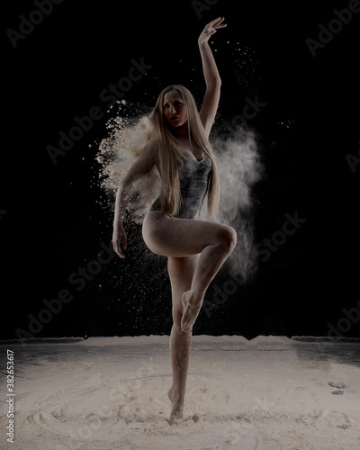 Beautiful sensual fit slim dancer in black body with long blond hair throwing dust, flour, powder on black background.