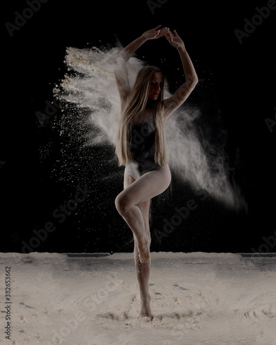 Beautiful sensual fit slim dancer in black body with long blond hair throwing dust, flour, powder on black background.