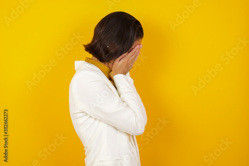 Sad young Caucasian woman wearing casual clothes standing against yellow background covering his face with hands and crying.