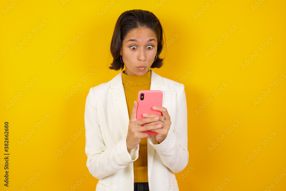 Horizontal shot of pretty young female isolated over pink background, looks with bugged eyes, holds modern smart phone, receives unexpected message from friend, reads reminder. Horizontal shot of pretty young female isolated over pink background, looks with bugged eyes, holds modern smart phone, receives unexpected message from friend, reads reminder.