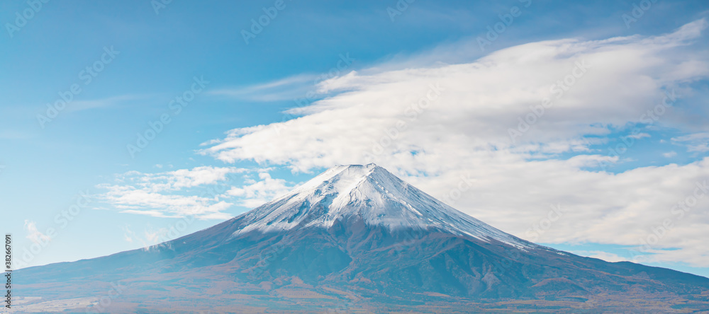 High angle view of Mount Fuji with snow covering the top of the volcano peak on a sunny day of autumn, Japan. There is a bright blue sky with a fluffy white cloud in a soft tone. There is copy space.