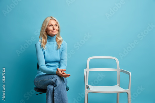 Photo of charming beaituful senior woman sits on comfortable chair enjoys good day at home concentrated above thoughtfully isolated over blue background makes plans for future. Elderly lifestyle photo