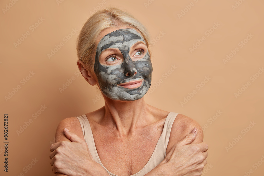 Portrait of thoughtful senior Caucasian woman crosses arms and touches shoulders gently wears homemade facial mask at home concentrated away isolated on beige background. Natural beauty concept Portrait of thoughtful senior Caucasian woman crosses arms and touches shoulders gently wears homemade facial mask at home concentrated away isolated on beige background. Natural beauty concept