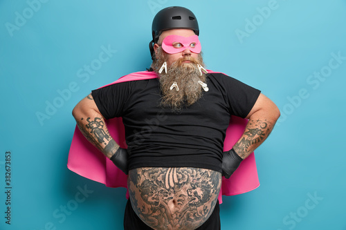 Photo of thoughtful bearded European man in superhero costume keeps hands on waist looks away and thinks deeply about something stands with tattooed fat belly against blue studio background.