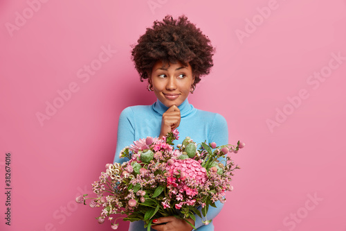 Portrait of thoughtful young Afro American woman touches chin holds beautiful bouquet of flowers in hands going to congratulate friend with special occasion isolated on pink background enjoys holiday