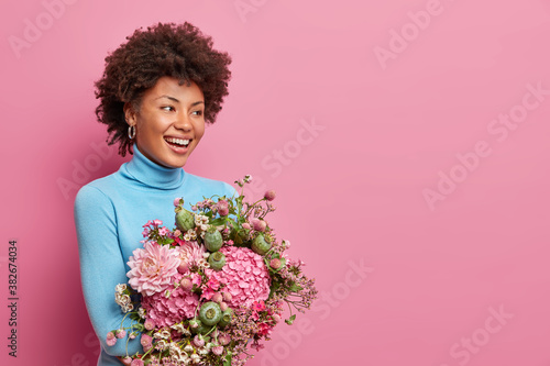 Half length shot of nice attractive Afro American woman enjoys festal event date occasion holds bunch of fresh flowers smiles broadly being in good mood poses against rosy wall with copy space