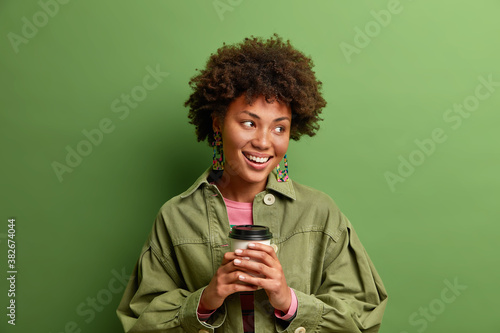 Delighted dark skinned female model with Afro hair looks away has toothy smile being deep in thoughts while drinks coffee wears stylish jacket isolated on green background. Lifestyle and rest