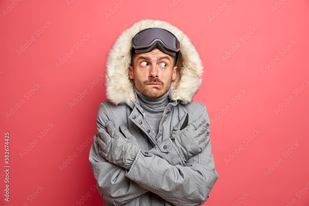 Puzzled frozen man crosses hands and tries to warm himself trembles from cold during snowy low temperature weather wears warm winter jacket gloves and ski goggles shaking freezing on frosty day Puzzled frozen man crosses hands and tries to warm himself trembles from cold during snowy low temperature weather wears warm winter jacket gloves and ski goggles shaking freezing on frosty day