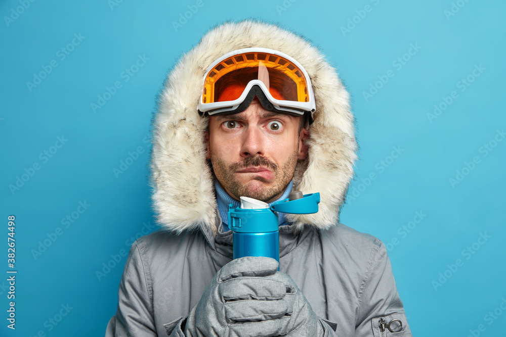 Serious man trembles from cold after going skiing during frosty winter day holds flask with hot beverage wears ski goggles and warm jacket isolated on blue background. Season recreation concept Serious man trembles from cold after going skiing during frosty winter day holds flask with hot beverage wears ski goggles and warm jacket isolated on blue background. Season recreation concept