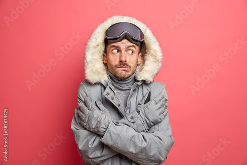 Puzzled frozen man crosses hands and tries to warm himself trembles from cold during snowy low temperature weather wears warm winter jacket gloves and ski goggles shaking freezing on frosty day