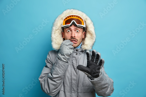 Scared emotional male skier wears warm jacket ski goggles and gloves stares shocked at camera has active winter holidays spends vacation in mountains isolated on blue background. Stunned snowboarder