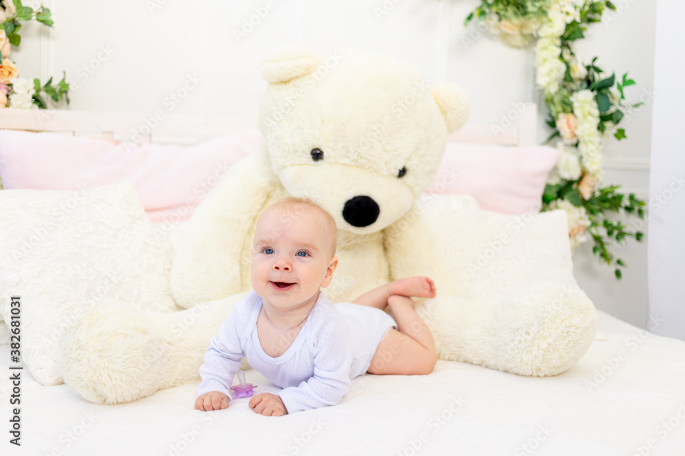 a small baby girl 6 months old is lying on a white bed at home with a large Teddy bear