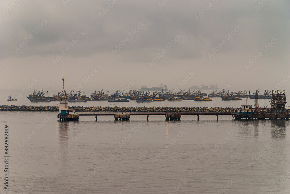 Lima, Peru - December 4, 2008: Early morning fog over fleet of fishing vessels moored on ocean water of Puerto Nuevo. Pier and protective dam up front.