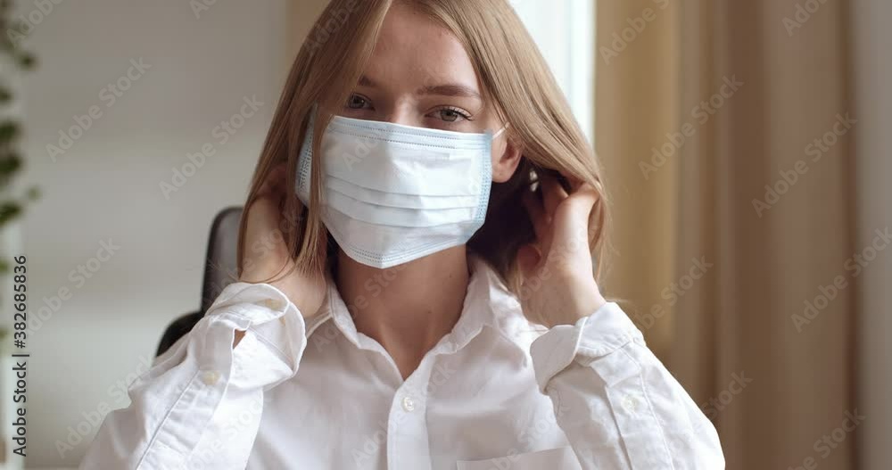 Portrait of young business woman attractive student sitting at office at table at home, looking at camera, putting on medical mask on female face protecting herself from coronavirus infection epidemic Portrait of young business woman attractive student sitting at office at table at home, looking at camera, putting on medical mask on female face protecting herself from coronavirus infection epidemic