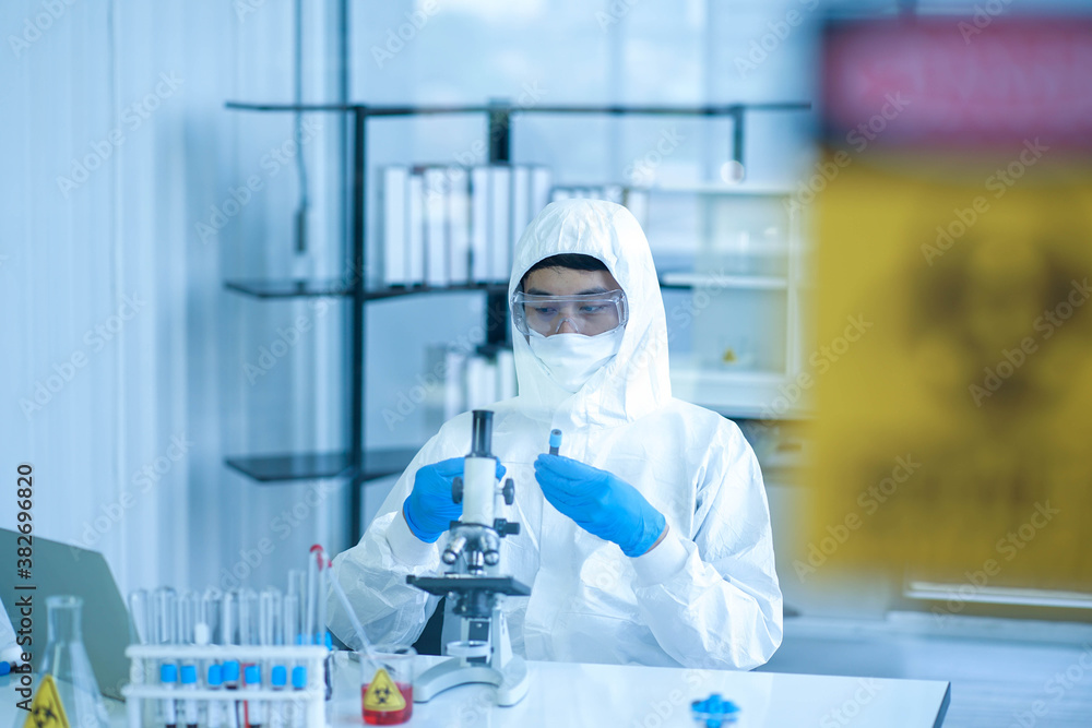 Fototapeta premium Asian medical scientist wearing protective white jumpsuit and face mask with glasses testing sample with microscope in clinical laboratory. Health care and medical concept. Virus pandemic concept. 
