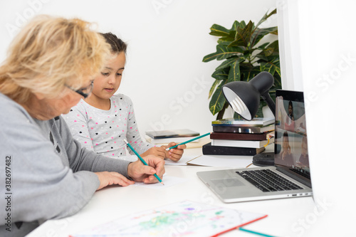 Cute and happy little girl child using laptop computer with her grandma, studying through online e-learning system.