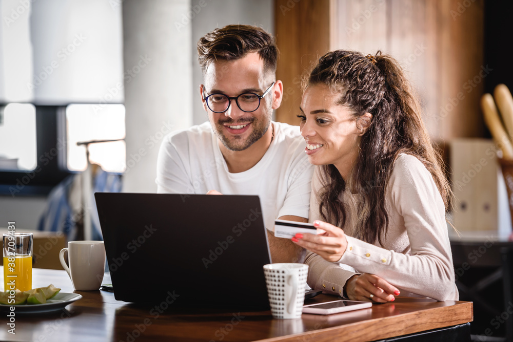 Smiling couple in kitchen ordering food online from restaurant. They using laptop and paying with credit card. 