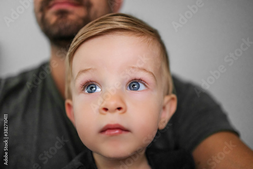 Youngster sitting in father's lap and looking at something with his beautiful big blue eyes.
