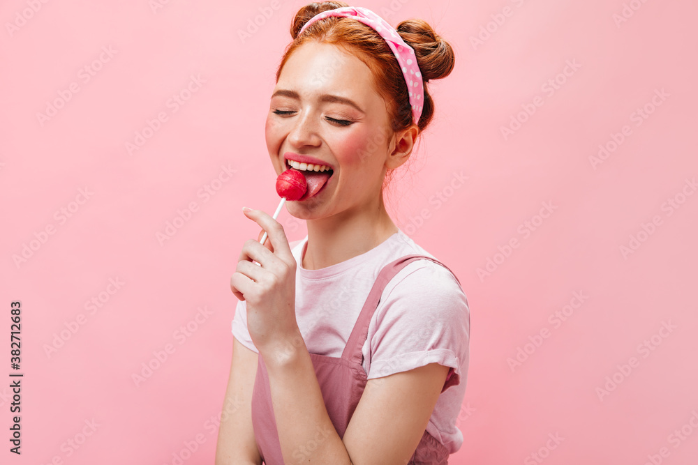 Cute redhead woman in white top and dress licks lollipop on pink background Cute redhead woman in white top and dress licks lollipop on pink background