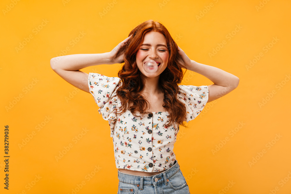 Positive curly girl in white cropped top touches her red curly hair and inflates bubble of gum Positive curly girl in white cropped top touches her red curly hair and inflates bubble of gum