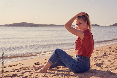 Happy and beautiful young white woman is sitting on the sandy beach in sunlight in blue jeans and red shirt and lookin back. Copy space.